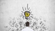 Rear view of the young manager, who is trying to find new idea. Business icons and an yellow light bulb are drawn on the concrete wall.